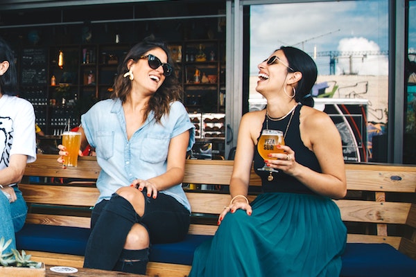 Two travelling solo women laugh and drink beer in a hostel bar.
